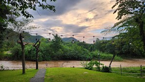A riverside pathway with a wooden bridge, surrounded by lush greenery and a mountainous backdrop. - Seven 50s (Pai)