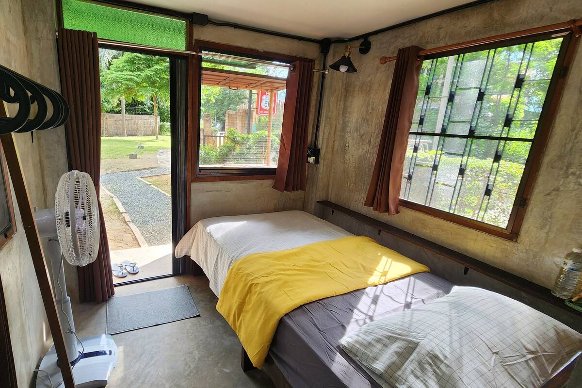 A small, compact room with a single bed, a fan, and a window with curtains.
