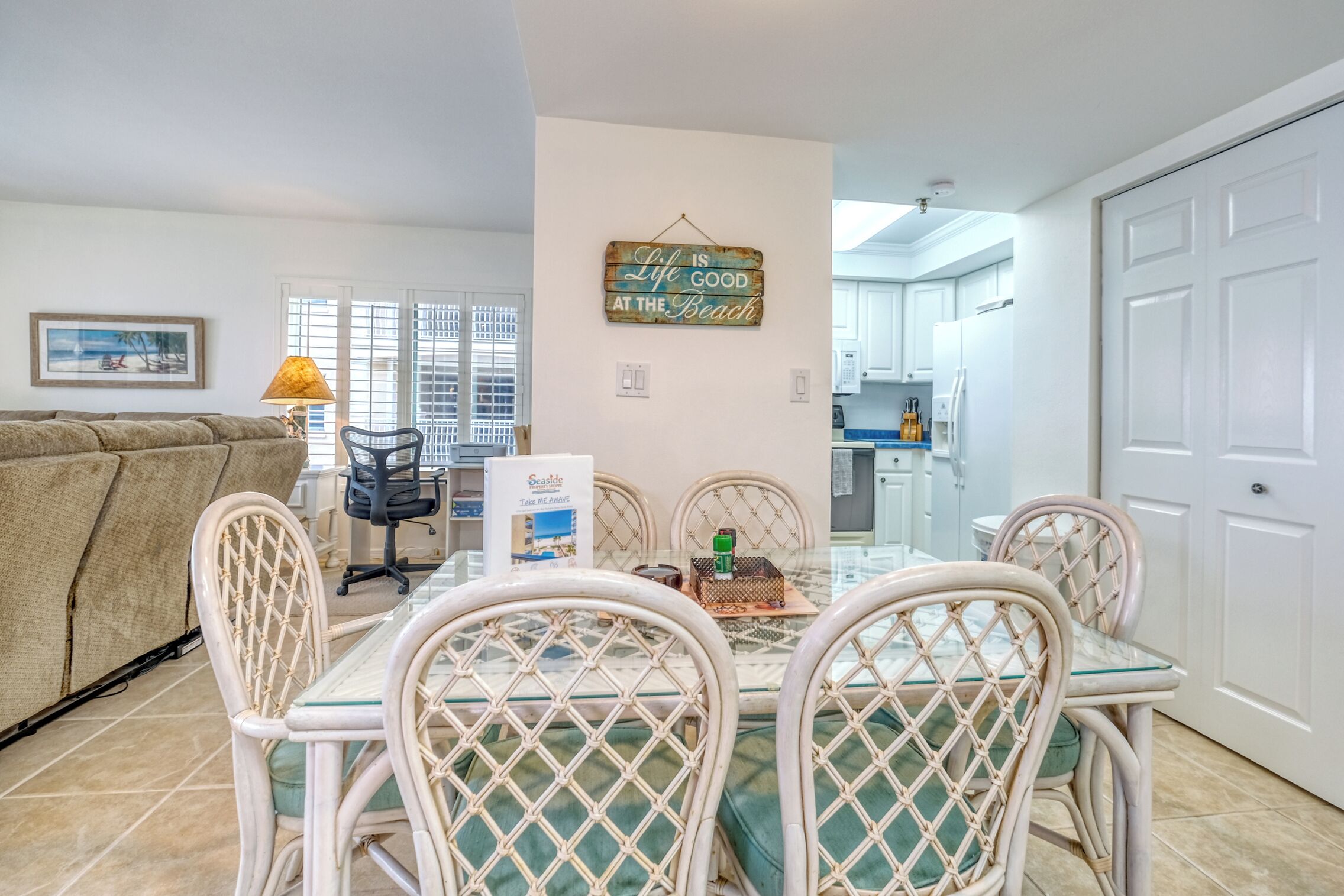 A dining area with a glass table and wicker chairs, a sofa, a lamp, and a kitchen in the background.