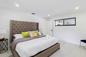 A modern bedroom with a tufted headboard, a large bed with white and grey bedding, a bedside table with a lamp, and a window with blinds. - Home 6 Mi to Hollywood Beach: Grill, Swim, Relax (Hollywood)