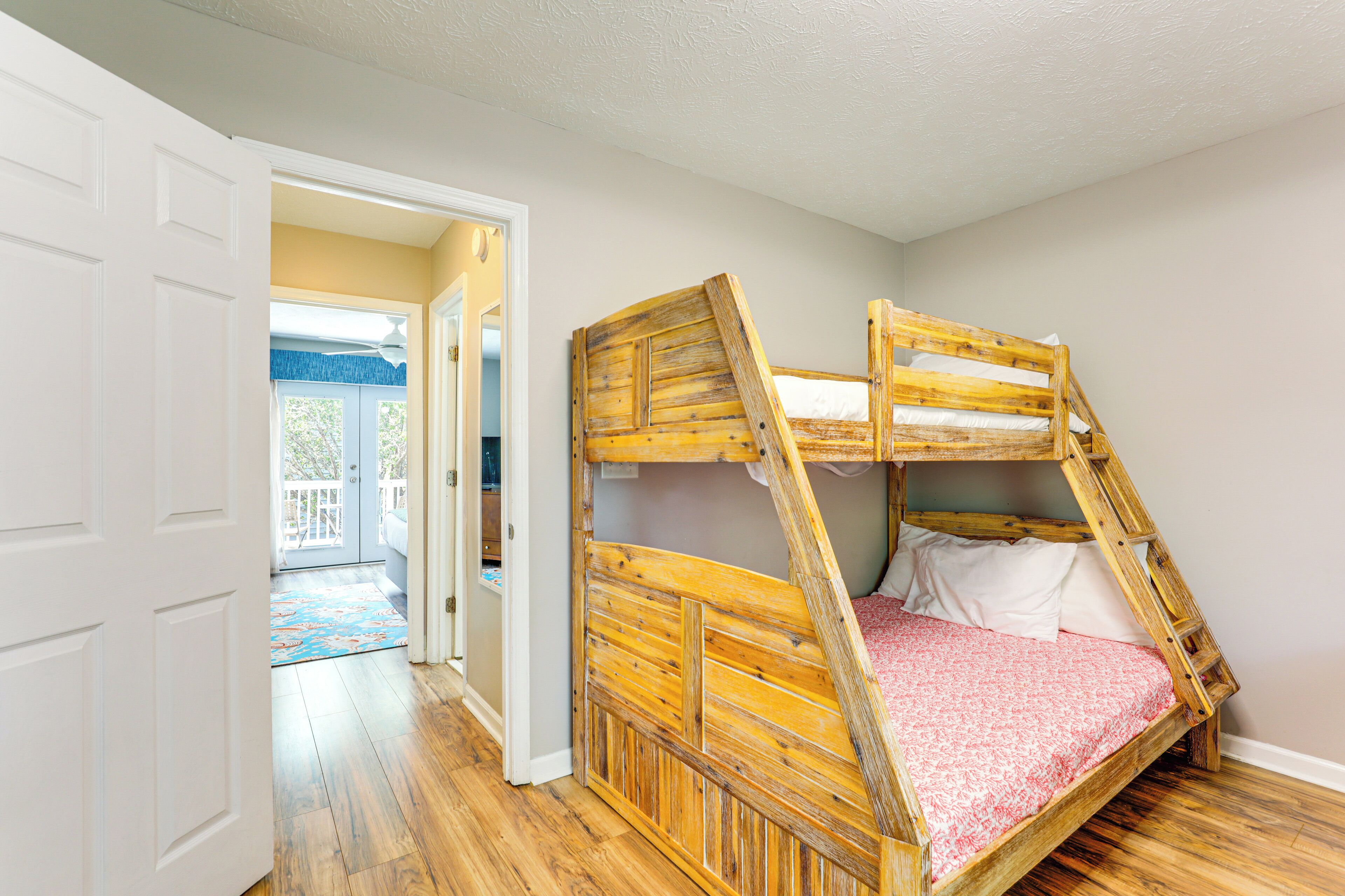 A room with a bunk bed, wooden flooring, a door leading to a balcony, and a view of the outdoors.