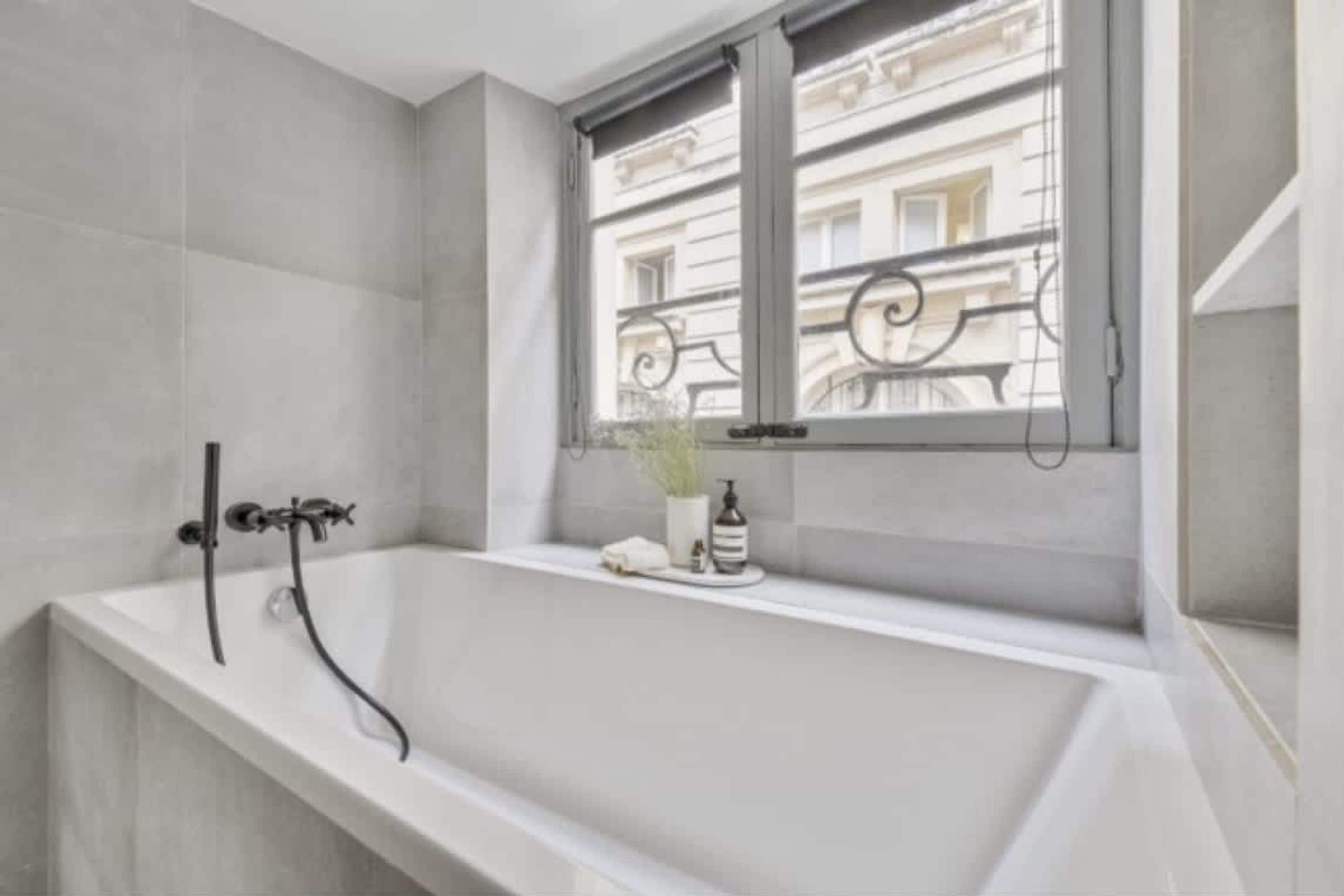 A modern bathroom with a freestanding bathtub, a window with a view of buildings, and a small plant on the windowsill.