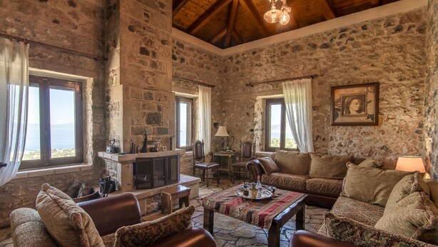 A rustic stone-walled room with a fireplace, leather sofas, and a view of the sea. - Niki s Tower - Majestic Seaview Stone Nest (Kalamata)