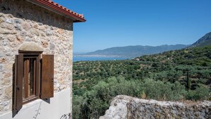 A stone building with a red-tiled roof and a wooden door. It overlooks a mountainous landscape with dense vegetation and a distant view of a city. - Niki s Tower - Majestic Seaview Stone Nest (Kalamata)
