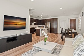 A modern living room with a flat-screen TV, a coffee table with a vase of flowers, a sofa, and a dining area with a table and chairs. - Comfort Style Home Near the Airport (Ontario)