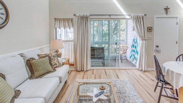 Una sala de estar con sofá, mesa de centro y comedor con mesa y sillas. Hay un ventanal corredera que da a un área exterior con una silla y una mesa.