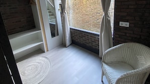 A modern room with a brick wall, a large window with curtains, a white wicker chair, and a built-in shelf unit. - Central apartment - perfect base for nature, culture and cities (Soest)