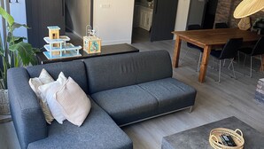 A modern living room with a grey sectional sofa, a wooden dining table with chairs, and a small side table with decorative items. - Central apartment - perfect base for nature, culture and cities (Soest)