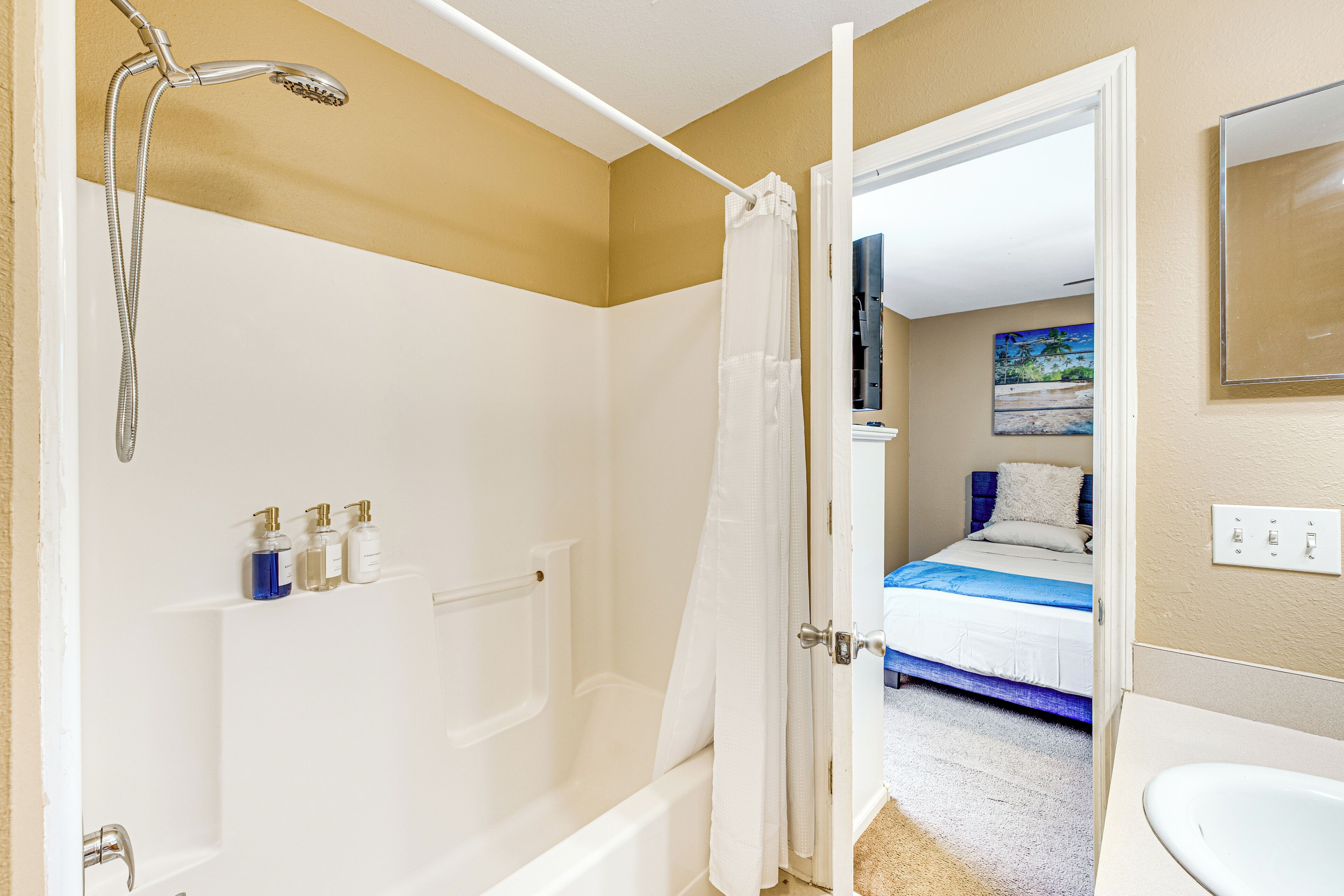 Baño con bañera, ducha e inodoro. Se ve una cama a través de una puerta abierta al fondo.