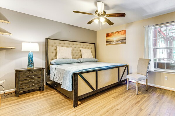 A bedroom with a bed, bedside table, lamp, chair, ceiling fan, window, and artwork.