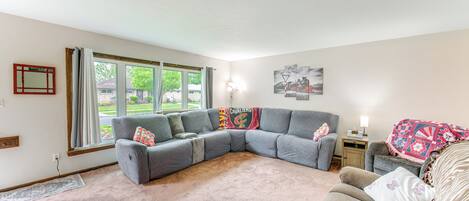 A living room with a gray sectional sofa, a brown couch, a rug, a window with curtains, a lamp, and a wall with a framed picture.