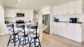 Una cocina moderna con muebles blancos, un refrigerador de acero inoxidable, microondas y una estación de café. Hay tres taburetes de bar con asientos acolchados.