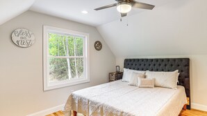 Une chambre avec un grand lit, un ventilateur de plafond, une fenêtre donnant sur les arbres et un mur orné d’une inscription : « Que notre foyer soit béni ».
