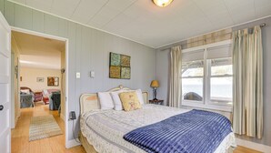 Un dormitorio con una cama grande, una mesita de noche con lámpara, una ventana con cortinas y una puerta que da a otra habitación.