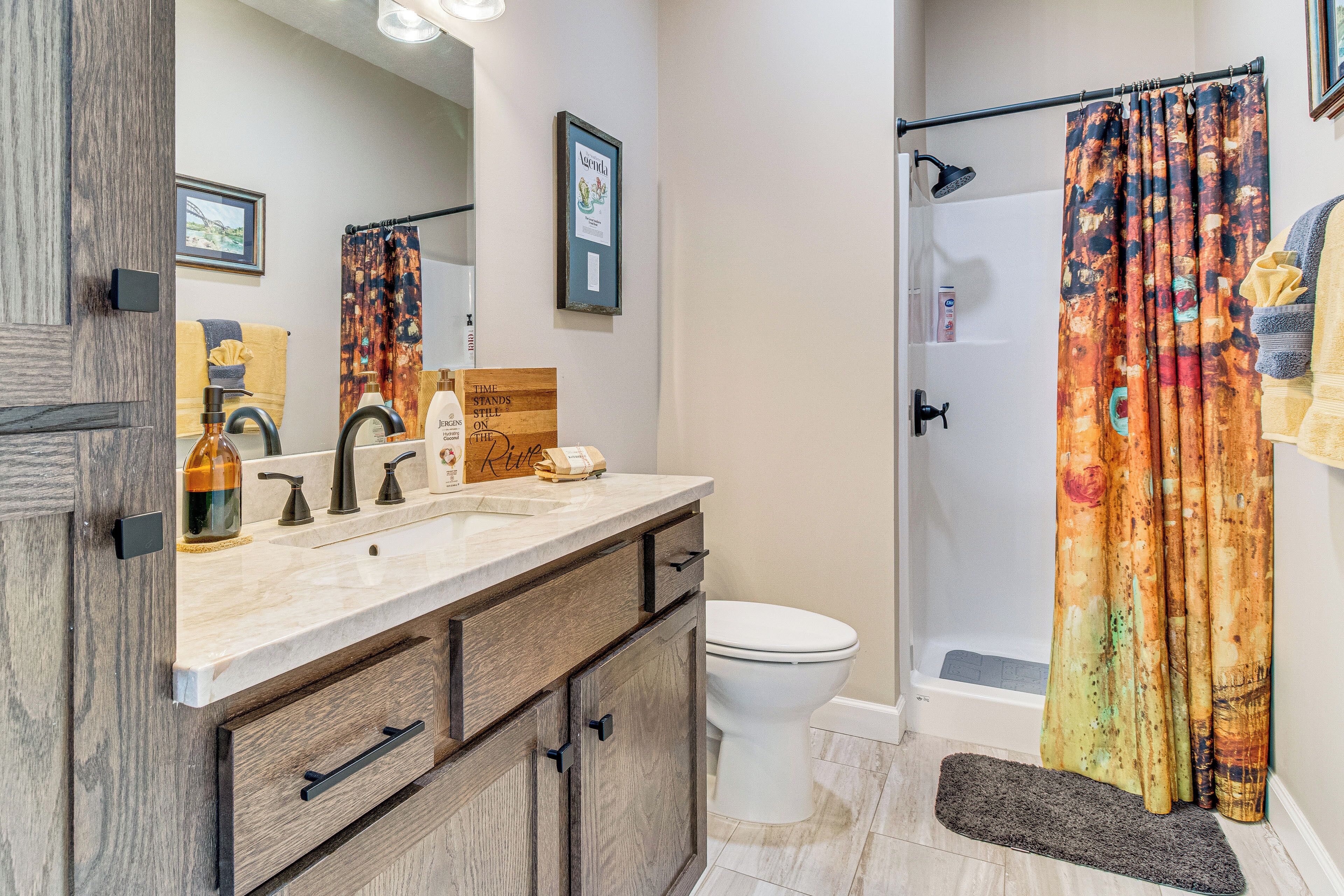 A bathroom with a wooden vanity, a white toilet, a shower with a colorful curtain, and a framed picture on the wall.