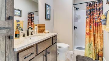 A bathroom with a wooden vanity, a white toilet, a shower with a colorful curtain, and a framed picture on the wall.