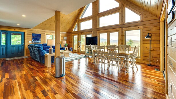 A spacious living room with wooden floors, a blue door, a sofa, a TV, and a dining area with chairs and a table. - Mtn-view Deck: Scenic Conway Log Cabin! (Conway)