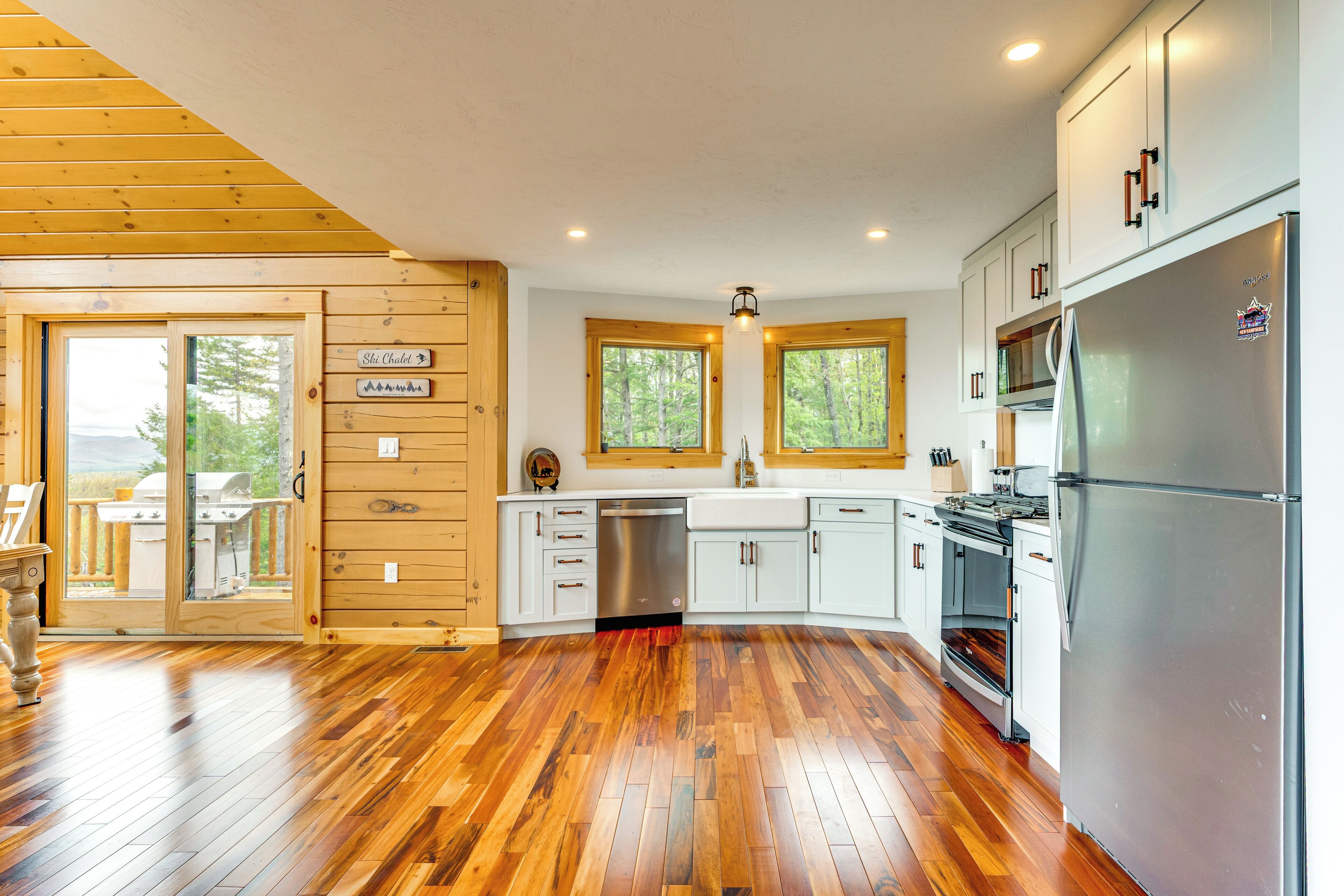 A modern kitchen with wooden flooring, white cabinets, stainless steel appliances, and a view of the outdoors through large windows.