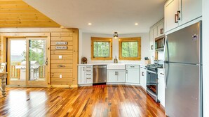 A modern kitchen with wooden flooring, white cabinets, stainless steel appliances, and a view of the outdoors through large windows. - Mtn-view Deck: Scenic Conway Log Cabin! (Conway)
