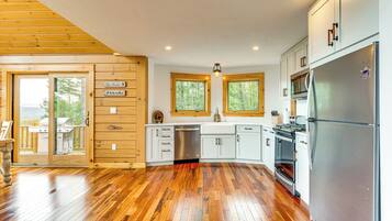 A modern kitchen with wooden flooring, white cabinets, stainless steel appliances, and a view of the outdoors through large windows.