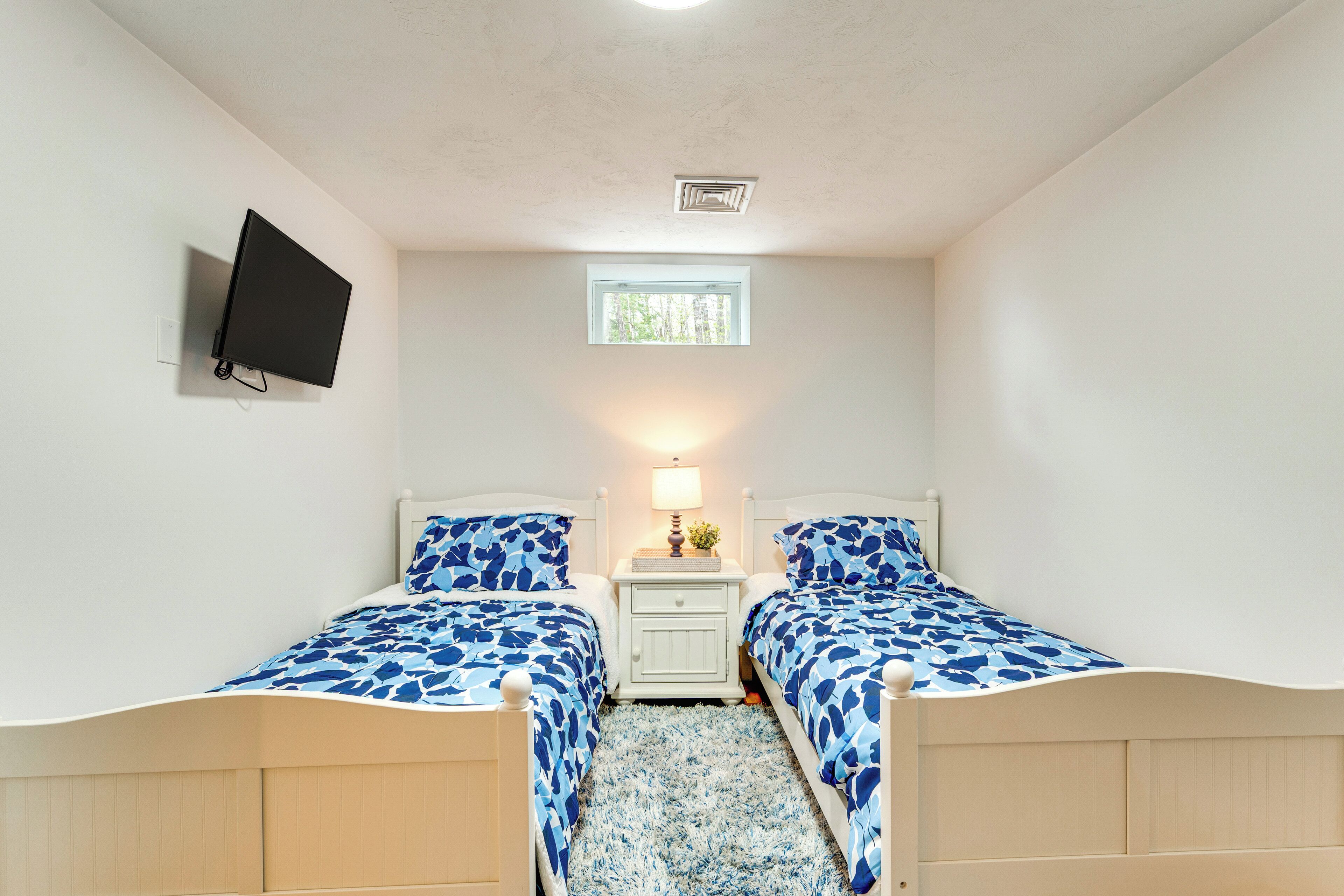 A bedroom with two beds, a TV mounted on the wall, a window, and a nightstand with a lamp.