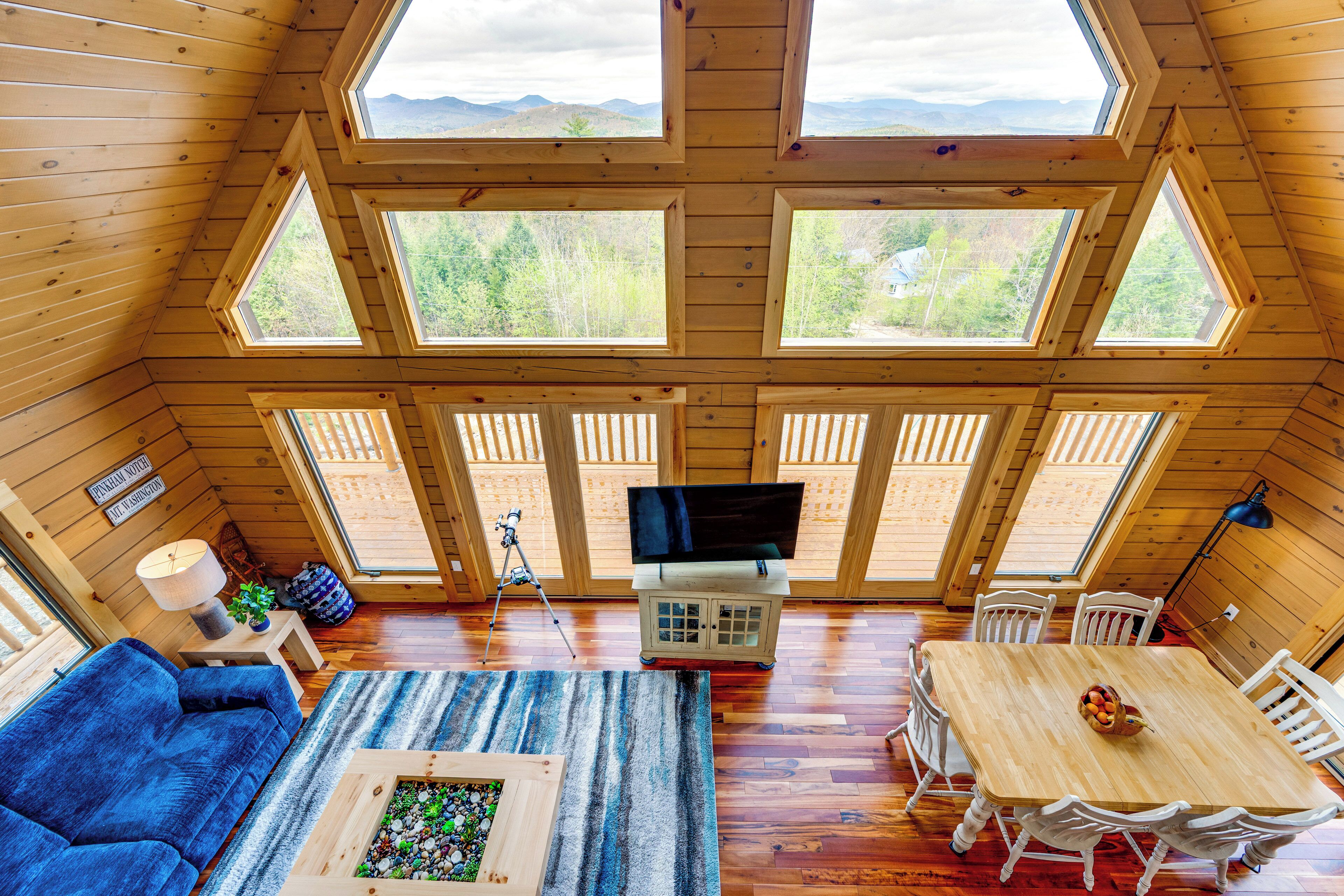 A cozy cabin with a large wooden ceiling, multiple windows offering views of the outdoors, a dining area with a table and chairs, and a living room with a sofa and a coffee table.