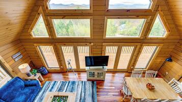 A cozy cabin with a large wooden ceiling, multiple windows offering views of the outdoors, a dining area with a table and chairs, and a living room with a sofa and a coffee table.