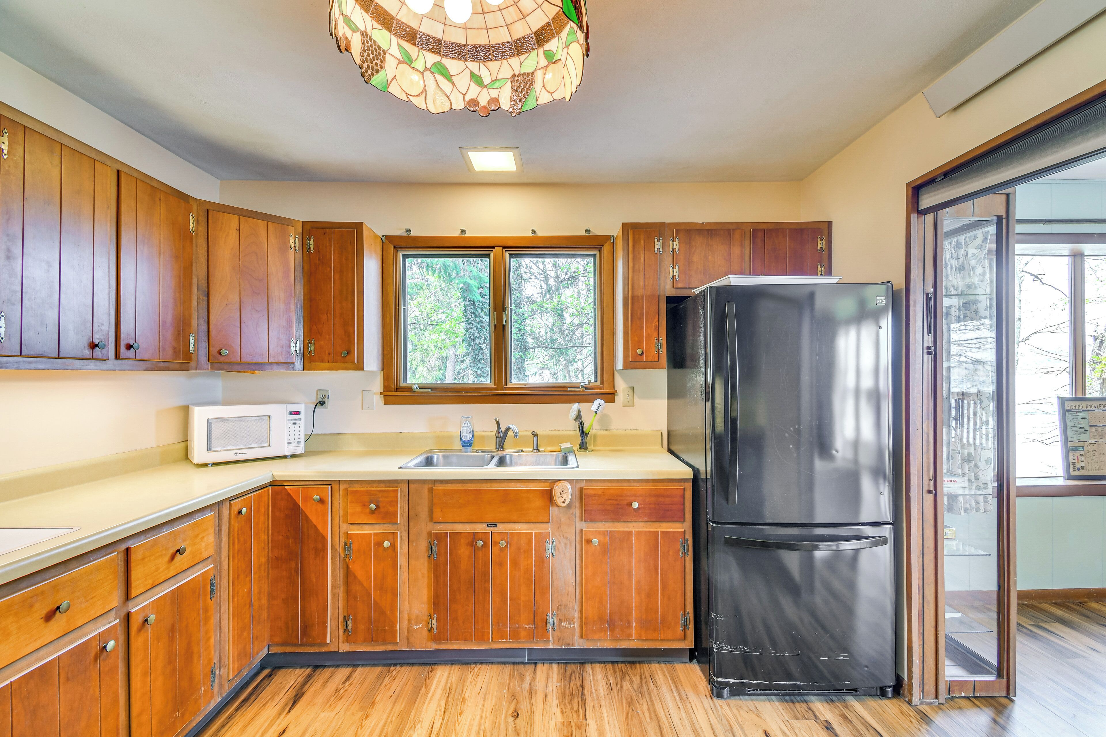 A kitchen with wooden cabinets, a stainless steel refrigerator, a microwave, and a window with a view of trees.