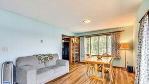 A room with a grey sofa, wooden dining table, and chairs. There is a heater, a window with curtains, and a door leading to another room. - < 1 Mi to State Park: Trumansburg Cottage w/ Deck! (Trumansburg)
