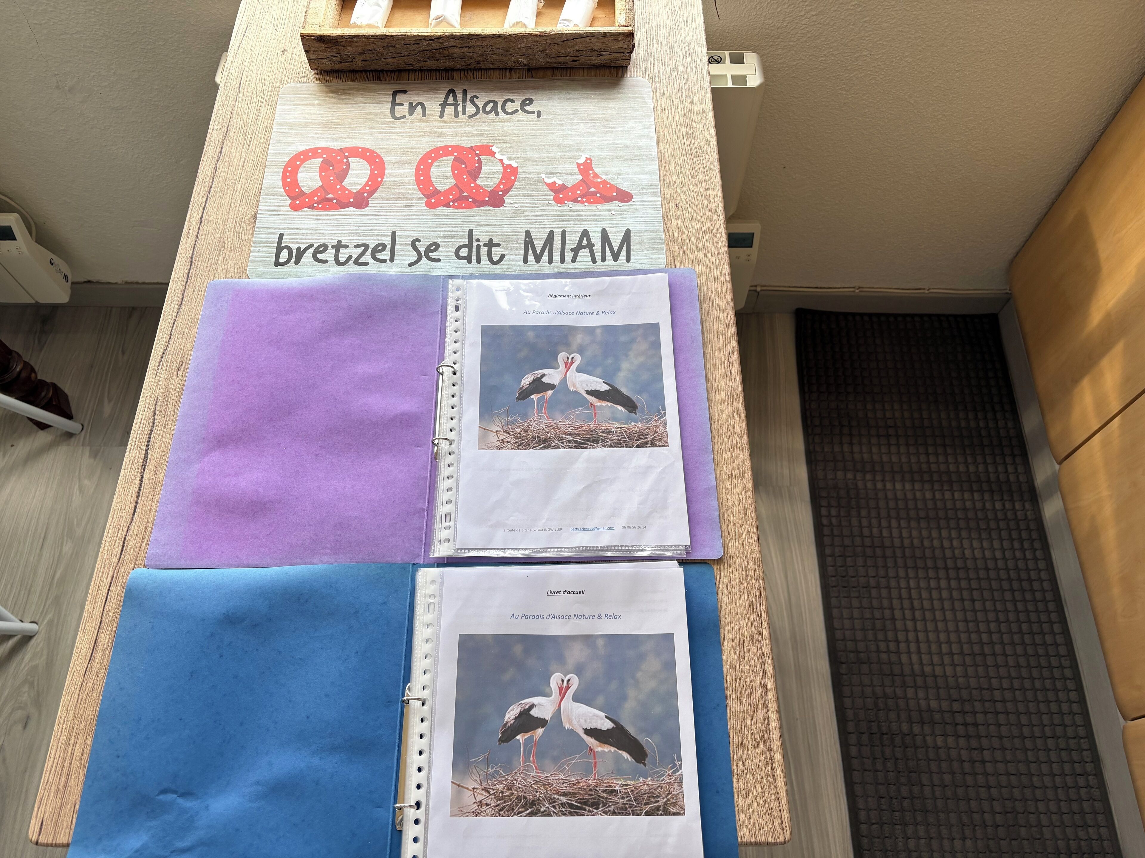 Ein hölzernes Schild mit Brezel-Illustrationen und Beschriftung, eine violette Ordner und ein blauer Ordner mit Vogelbildern, alles auf einer Holzoberfläche.