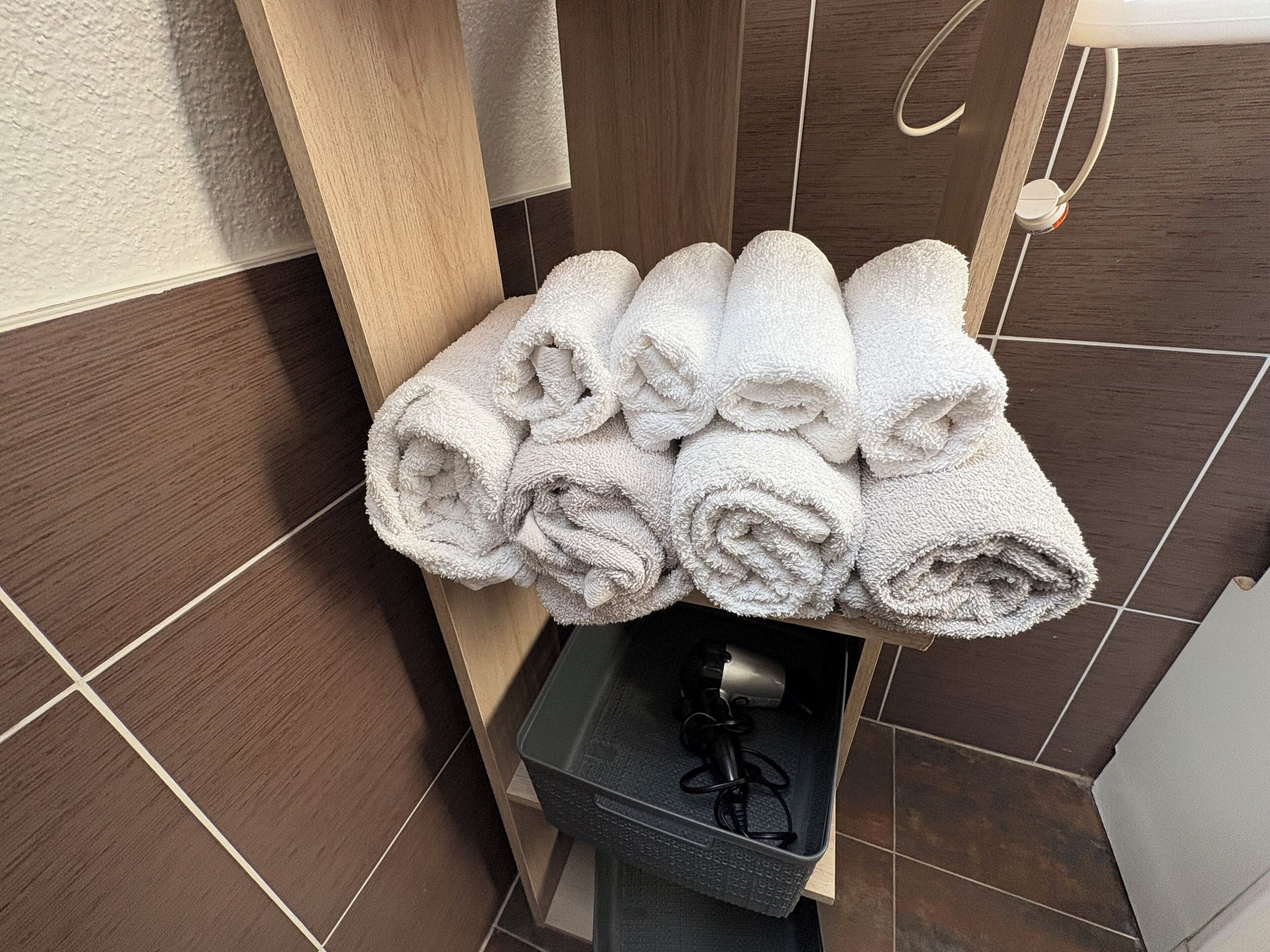 Un mucchio di asciugamani bianchi ordinatamente piegati su uno scaffale in un bagno con piastrelle marrone scuro.