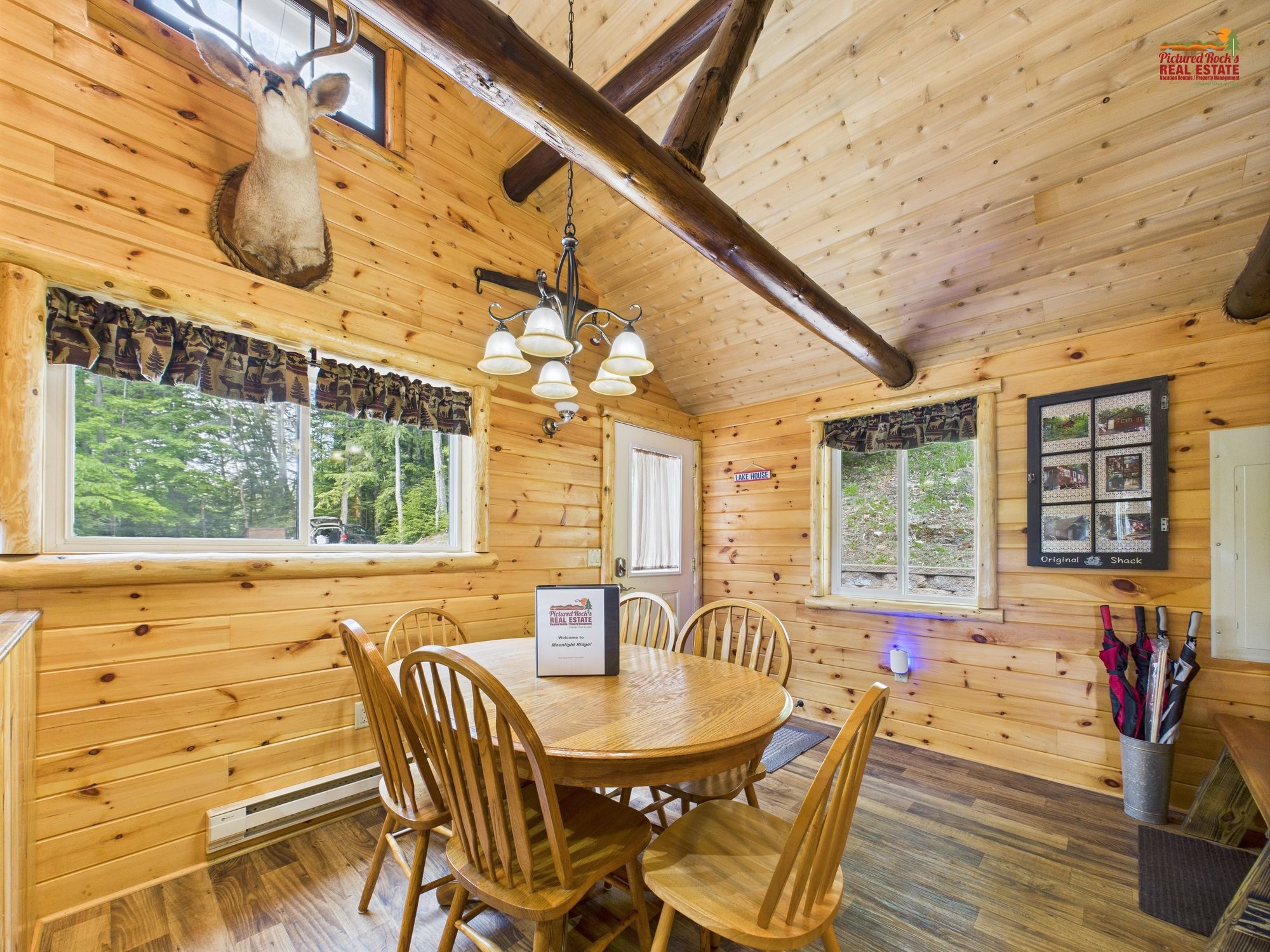 A wooden cabin interior with a dining table, chairs, a chandelier, and a mounted deer head.
