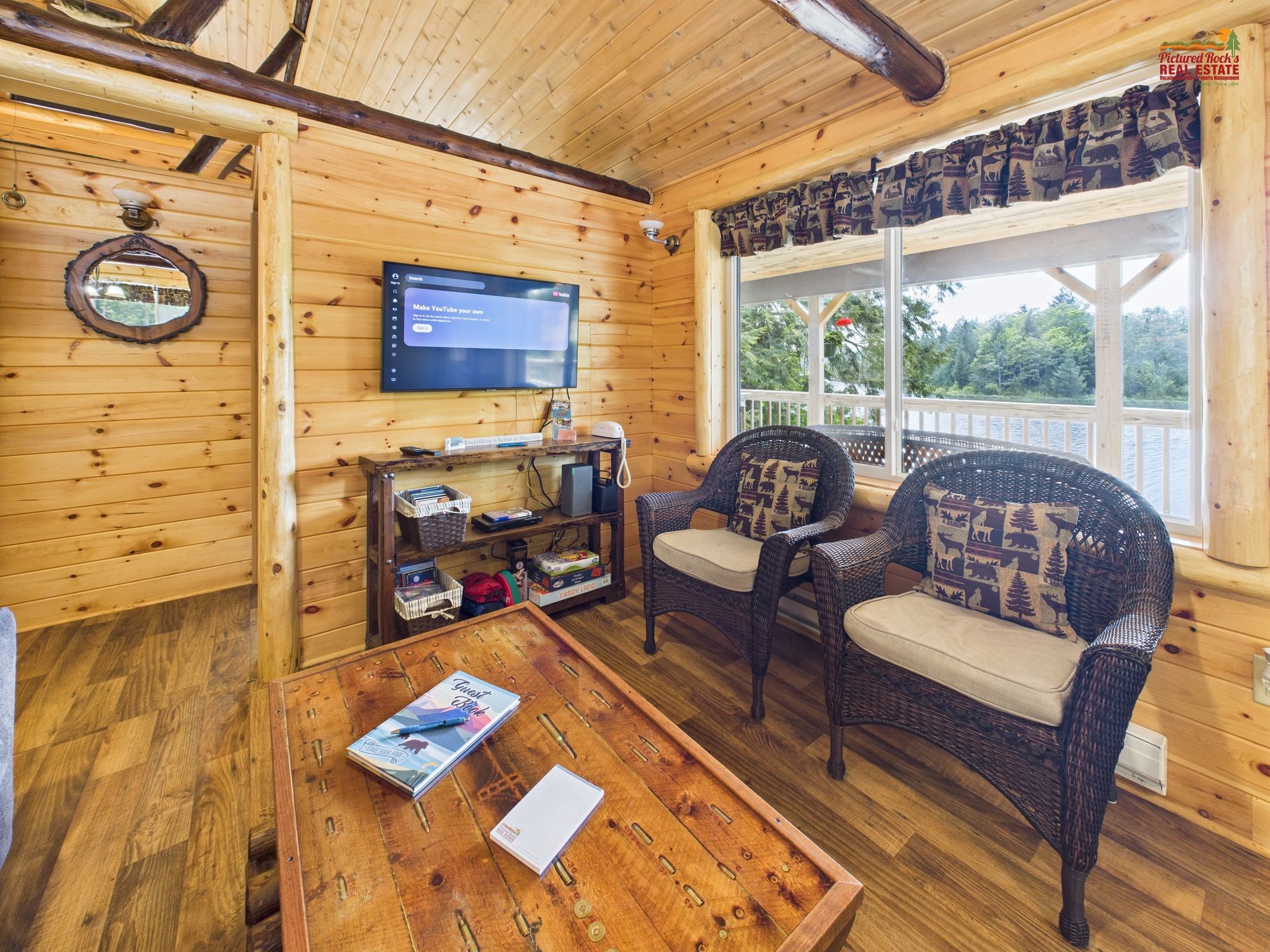 Interior acogedor de una cabaña con paredes de madera, un televisor, sillas de mimbre, una mesa de centro y un ventanal con vista a árboles.