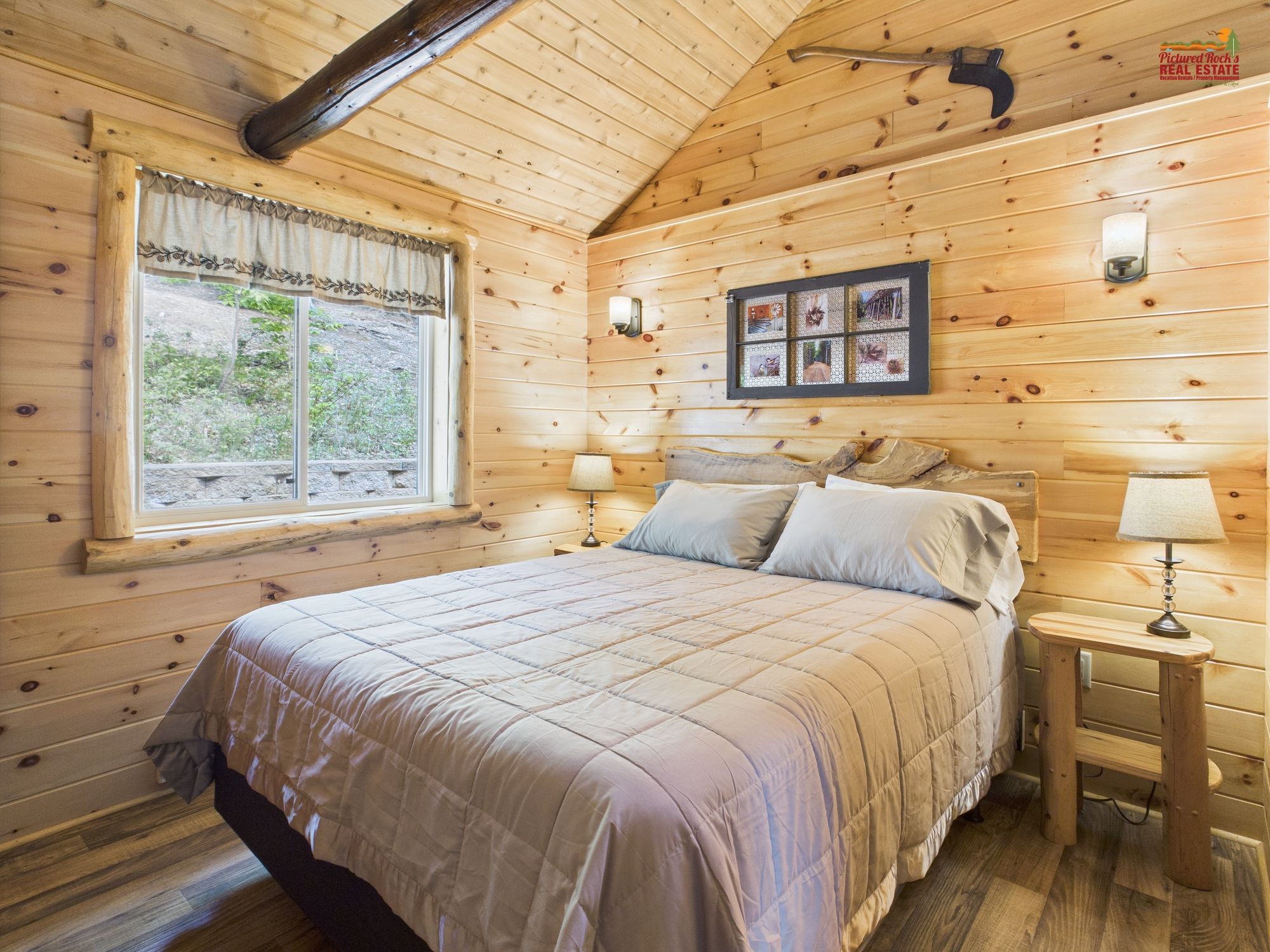 Un dormitorio acogedor tipo cabaña de madera, con una cama grande, una ventana con cortinas, una mesita de noche con una lámpara y un cuadro en la pared.