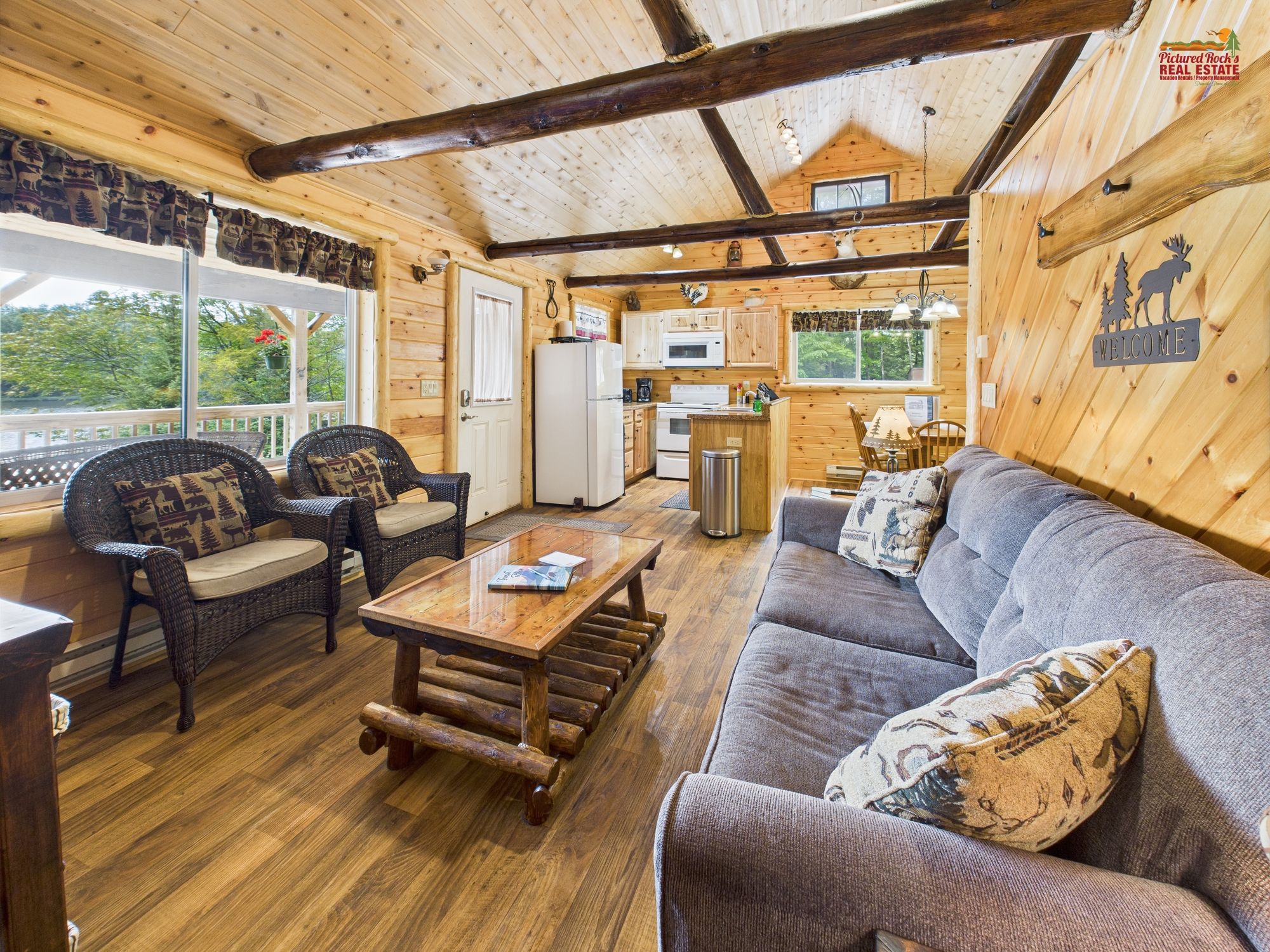 A cozy cabin interior with a wooden floor, a sofa, two armchairs, a coffee table, a kitchen area, and a large window offering a view of the outdoors.
