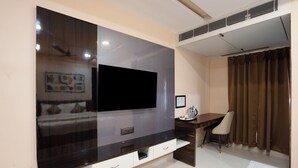 Un soggiorno moderno con un'ampia televisione a schermo piatto fissata a una parete in legno scuro, un mobile contenitore bianco con cassetti, una scrivania con sedia e una finestra con tende marroni.