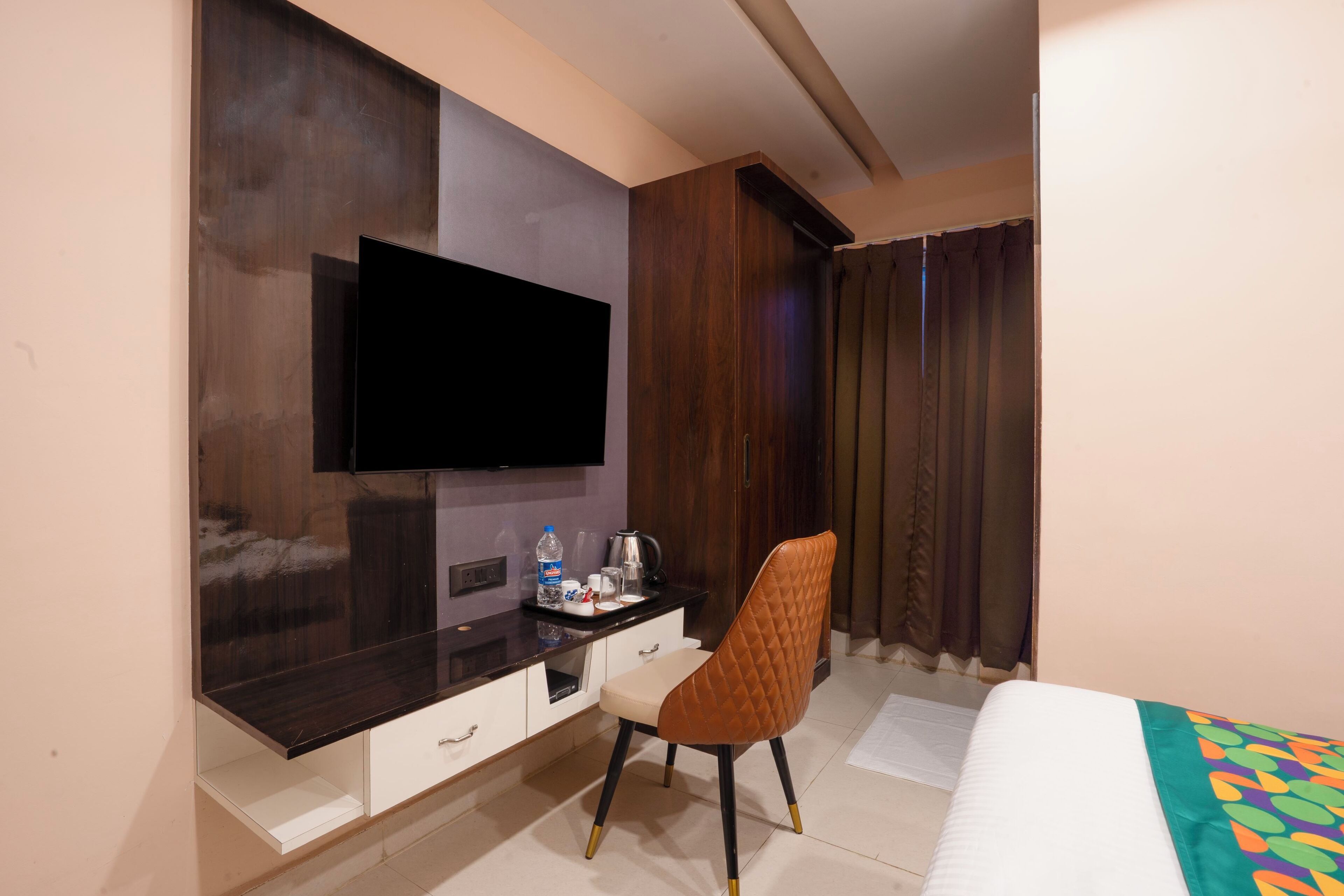 Une chambre d’hôtel dotée d’un téléviseur à écran plat fixé à un mur en bois foncé, d’un bureau blanc avec des tiroirs, d’une chaise rembourrée marron, d’un lit avec une couverture colorée et d’une fenêtre avec des rideaux.