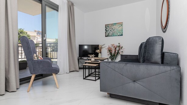 Un salon moderne avec un canapé gris, un fauteuil gris, un téléviseur à écran plat, une petite table sur laquelle est posé un vase de fleurs et une grande fenêtre avec des rideaux.