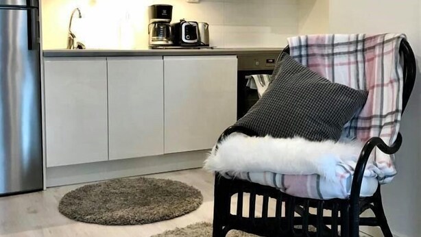 Ett modernt kök med vita skåp, ett kylskåp i rostfritt stål och en kaffebryggare på bänkskivan. Där står en svart stol med en rutig filt och en vit, fluffig kudde. Två runda mattor ligger på golvet.