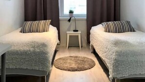 Ett litet, väl upplyst rum med två enkelsängar, ett litet bord och ett fönster med gardiner.
