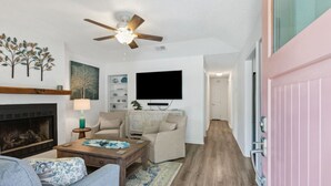 Un salon avec une cheminée, un ventilateur de plafond, un téléviseur et une porte rose.