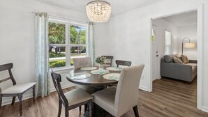 Un coin repas avec une table ronde, des chaises et un lustre. Une fenêtre avec des rideaux offre une vue sur la verdure à l’extérieur.