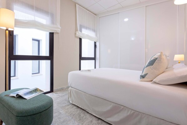 A hotel room with a bed, a green ottoman, and two windows with sheer curtains.