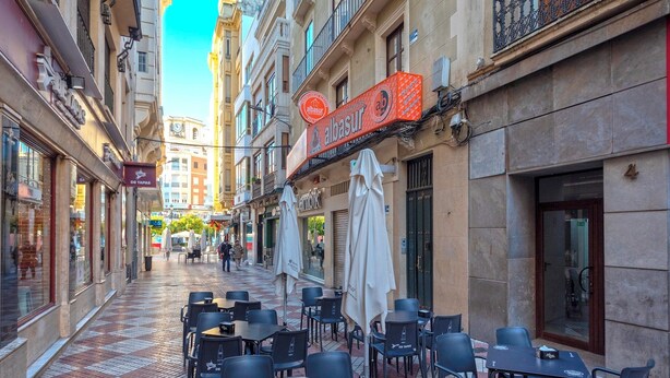 A narrow street with outdoor seating, a clock tower, and a building with a sign that reads "albasur".