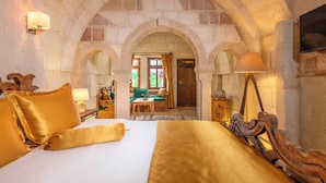 Habitación de hotel con una cama grande, dos mesitas de noche, una silla de madera y un arco de piedra que conduce a una zona de estar con una mesa y sillas.