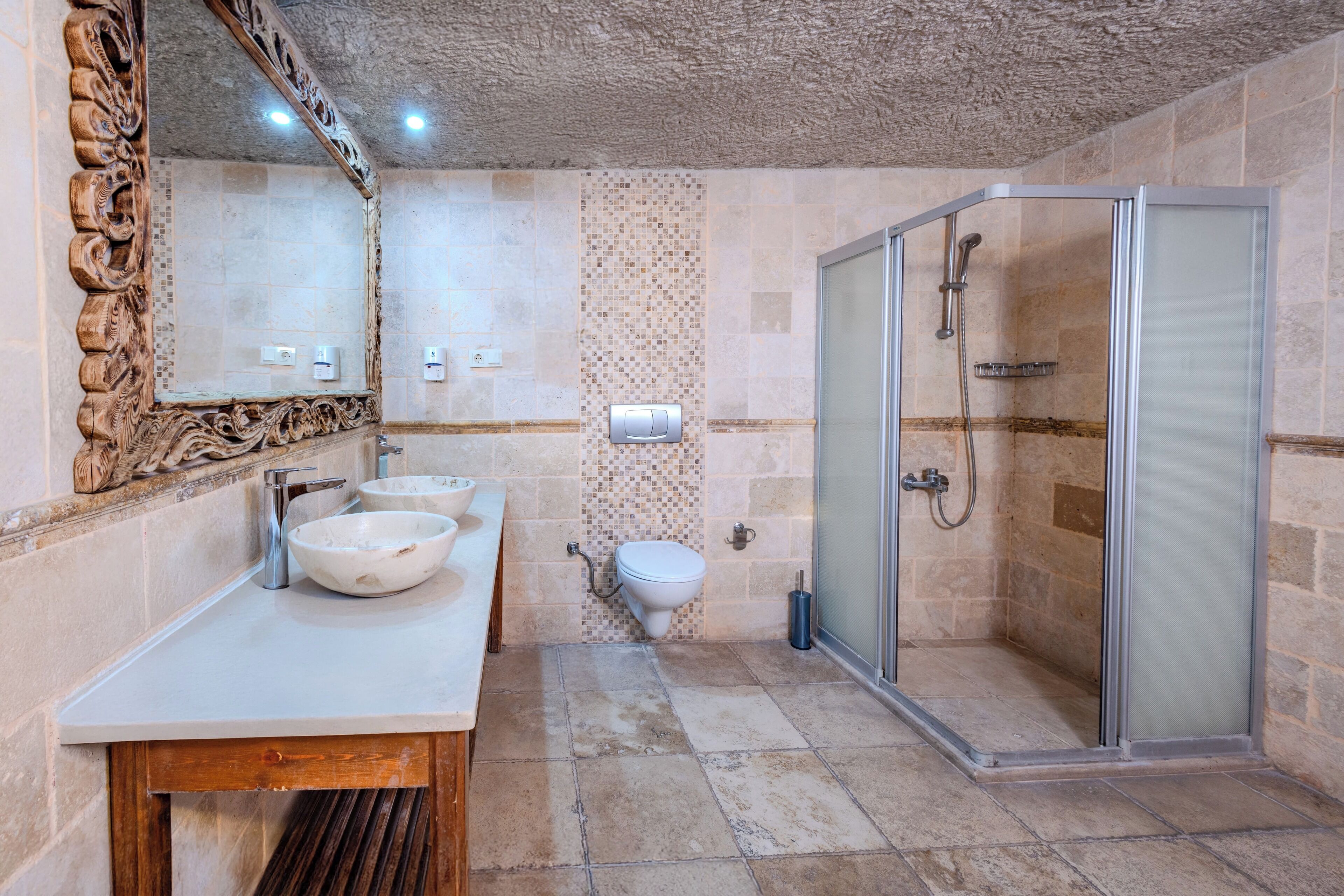 A bathroom with a wooden vanity, two sinks, a toilet, and a glass-enclosed shower.