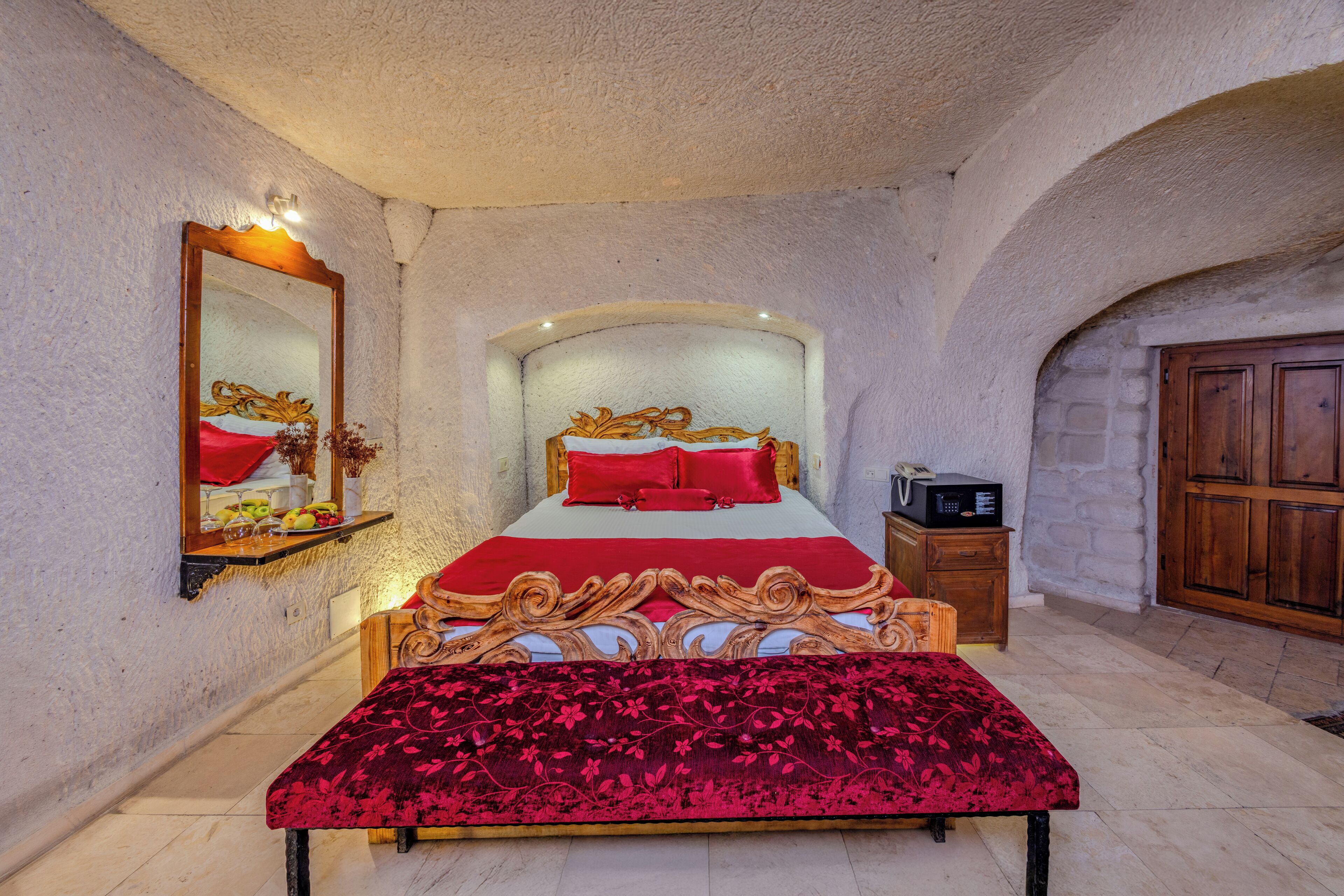 Un dormitorio con una cama grande, cabecera de madera, colcha roja, mesita de noche de madera, un espejo y una pared de piedra.