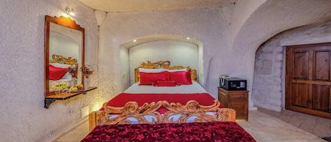 Un dormitorio con una cama grande, cabecera de madera, colcha roja, mesita de noche de madera, un espejo y una pared de piedra.