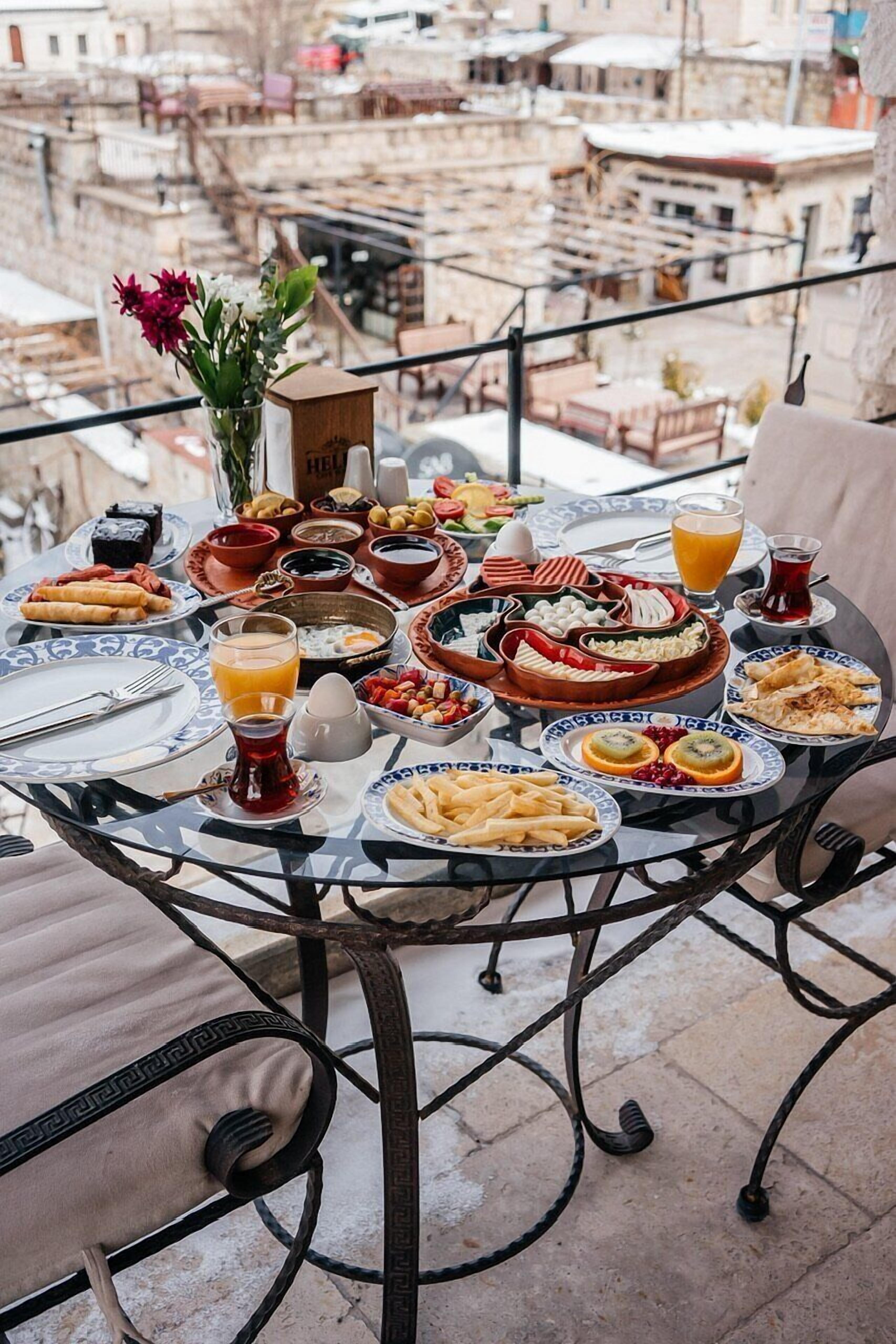 Une table dressée avec une variété de plats, notamment du pain, des œufs et des fruits, sur un balcon extérieur offrant une vue sur les bâtiments.