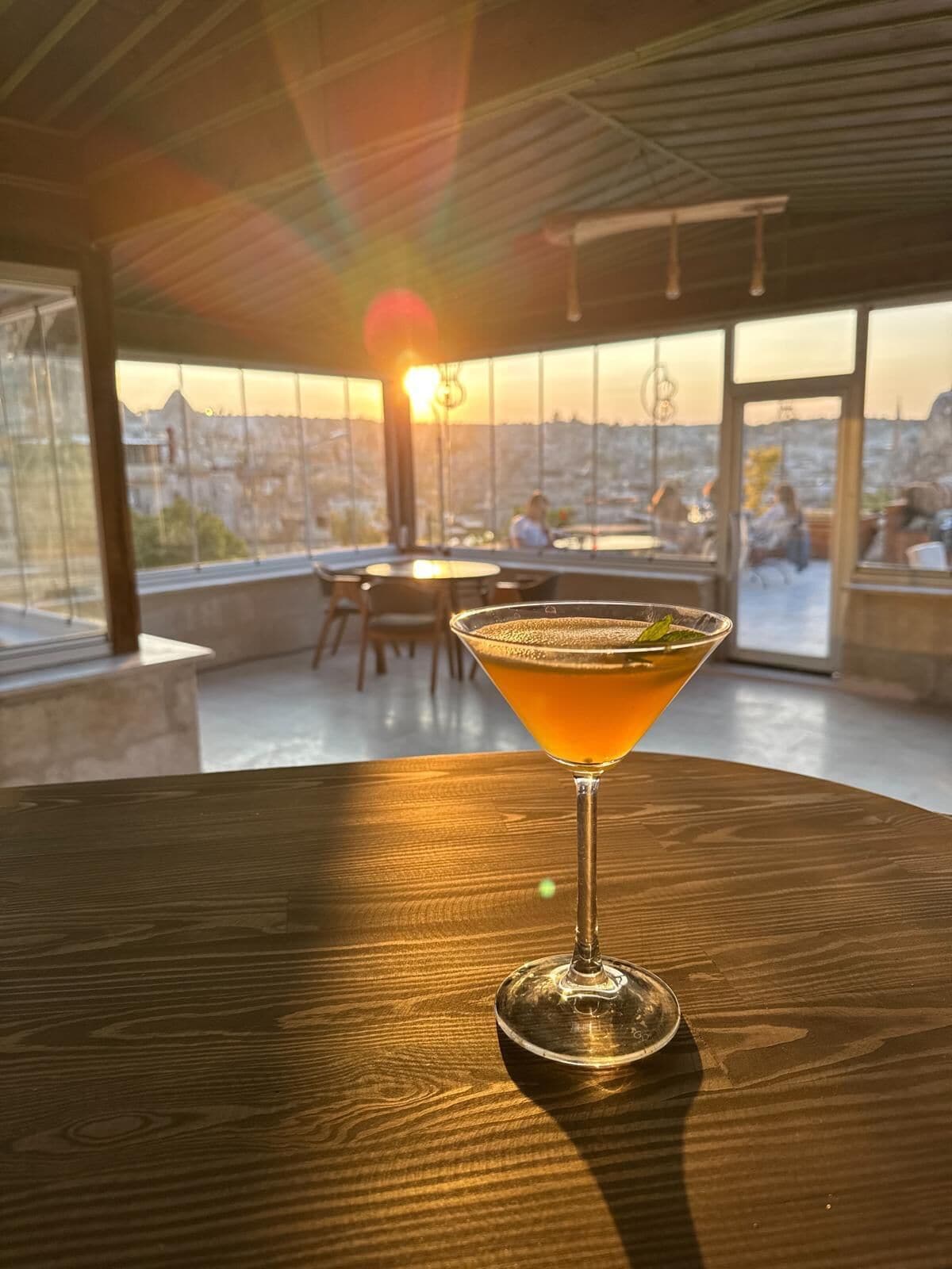 Un verre à martini posé sur une table en bois, avec une vue sur le coucher de soleil à travers de grandes fenêtres.